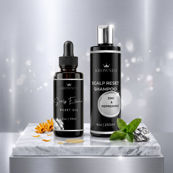Two KROWNED hair care products displayed on a white marble slab: a black dropper bottle labeled Scalp Elixir Reset Oil and a black pump bottle labeled Scalp Reset Shampoo with Zinc & Peppermint. The scene is styled with mint leaves, a small orange flower, and metallic accents against a soft silver bokeh background.